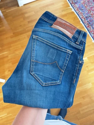 Jacob Cohën - Snygga blå  jeans från Jacob Cohën med klassisk femficksdesign och kontrastsömmar. Jeansen har en brun läderpatch med logga bak i midjan och är tillverkade i stretchigt denimtyg för extra komfort. Jeansen är i storlek 31. Uppsydda och sitter som 31/32. Modell Nick
