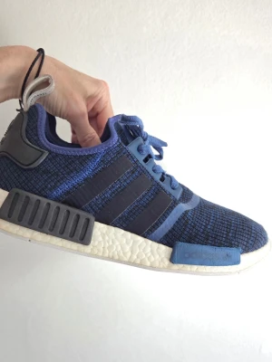 Blå Adidas NMD R1 sneakers - Säljer ett par blå Adidas NMD R1 sneakers med stickad ovandel, svarta detaljer och vita Boost-sulor. Skorna har de klassiska tre ränderna på sidan, svarta och blå block på mellansulan samt dragflik bak. Perfekta för en streetwear-look.