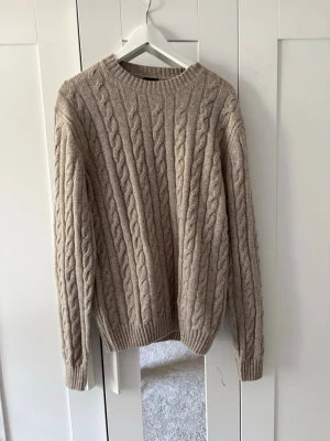 J.lindberg stickad tröja - Säljer en beige kabelstickad tröja med rund hals och långa ärmar. Tröjan har ett klassiskt flätmönster och ribbade muddar vid ärmslut och nederkant. Perfekt för dig som gillar en stilren och mysig look.