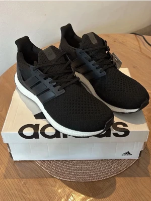 Svarta Adidas Ultraboost sneakers - Svarta Adidas Ultraboost sneakers med ventilerande mesh-överdel, vita Boost-mellansulor och svarta yttersulor med Continental-gummi. Klassisk tre-randig design på sidorna och snörning framtill. Perfekta för dig som gillar stilrena och bekväma sneakers.