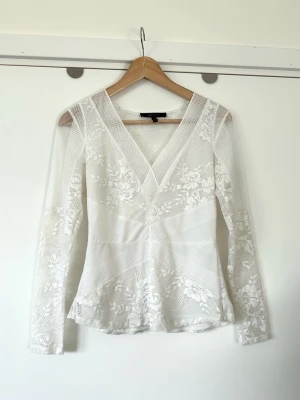 BCBGMAXAZRIA - vit v-ringad blus i spets - storlek XS - Stilren vit spetsblus från BCBG Max Azria i storlek XS. Tidlös modell med långa ärmar och V-ringning. Blusen har en figurnära passform med en lätt peplum-effekt nedtill, vilket ger en smickrande silhuett. Spetsmönstret varierar med blommiga detaljer och geometriska inslag, vilket skapar en dynamisk och elegant look. Ärmarna är också i spets med liknande mönster. Perfekt för både vardag och fest. Mycket gott skick!
