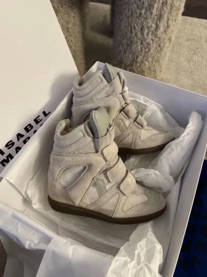 Beige sneakers med kilklack Isabel Marant - Säljer ett par ikoniska beige sneakers från Isabel Marant med kilklack och coola paneldetaljer. Skorna har kardborreband och är gjorda i mjuk mocka med gummisula. Perfekta för dig som vill ha både stil och höjd i vardagen.