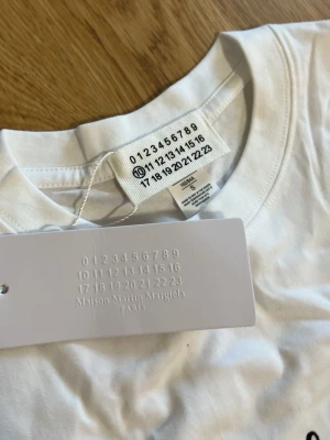 Vit Maison Margiela t-shirt med tryck - Vit t-shirt från Maison Martin Margiela med svart tryck av siffror och en markerad sexa på bröstet. Klassisk rund hals och korta ärmar. Tillverkad i mjuk bomull för en skön känsla och clean look.