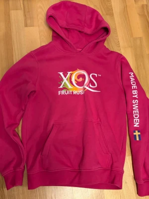 Rosa XQS Fruit Rush hoodie - Snygg rosa hoodie med XQS Fruit Rush-tryck på bröstet och texten 'MADE BY SWEDEN' samt svensk flagga på ena ärmen. Stor känguruficka framtill och huva. Materialet ser ut att vara mjuk bomullsblandning, perfekt för chill dagar.