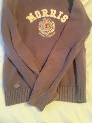 Mörkblå stickad tröja från Morris - Säljer en l stickad tröja från Morris med tryckt logga och emblem på bröstet. Tröjan har ribbade muddar vid ärmslut och nederkant samt rund halsringning. Perfekt för dig som gillar klassisk college-stil.