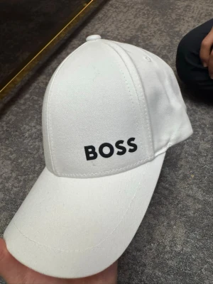 Vit keps från BOSS - Snygg vit keps från BOSS med klassisk böjd skärm och svart BOSS-logga broderad framtill. Tillverkad i ett slitstarkt tyg med justerbar rem bak för perfekt passform. Enkel och stilren design som passar till det mesta.