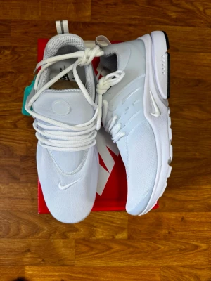 Nike Air Presto vita sneakers - Nike Air Presto sneakers i helvitt med mesh- och syntetmaterial. Skorna har en clean design med vita snören, diskret swoosh-logga på tå och sidor samt en bekväm, låg siluett. Perfekta för dig som gillar stilrena och sportiga sneakers.