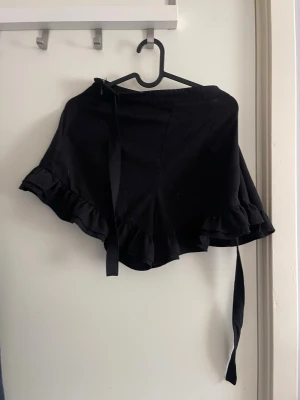 Svarta omlottkjol med volang - Supersnygg svart omlottkjol med volangkant och knytband i midjan. Kjolen har en flowy passform och är perfekt för dig som gillar en feminin och trendig stil. Tillverkad i mjukt tyg som känns skönt mot huden.
