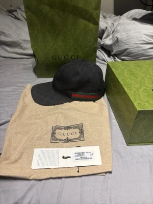 Svart keps från Gucci med GG-mönster - Snygg svart keps från Gucci med klassiskt GG-monogram över hela kepsen. På sidan finns den ikoniska gröna och röda randen. Kepsen har böjd skärm och justerbar rem bak. Materialet är canvas med detaljer i läder.