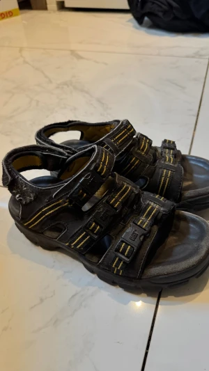 Svarta sportiga sandaler med gula detaljer - Säljer ett par svarta sandaler med gula sömmar och justerbara kardborreband. De har en sportig look med robust sula och flera remmar för bra passform. Perfekta för dig som gillar en aktiv stil och vill ha bekväma skor till sommaren.