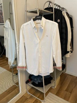 Vit skjorta med färgdetaljer från Burberry  - Klassisk vit skjorta i bomull med långa ärmar och knappar framtill. Ärmsluten har snygga ränder i rött och beige som ger en unik touch. Perfekt till jeans eller chinos för en clean och stilren look.