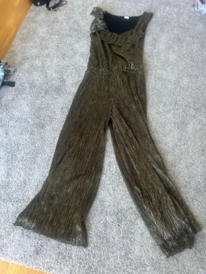 Glittrig plisserad jumpsuit i brons - Snygg jumpsuit i glittrigt bronsfärgat tyg med plisserad struktur. Asymmetrisk volang upptill och vida ben ger en trendig look. Perfekt för dig som vill sticka ut! Materialet känns lätt och följsamt.