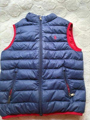 Blå dunväst från Polo Ralph Lauren - Snygg blå dunväst från Polo Ralph Lauren med röd insida och röda detaljer. Västen har två dragkedjeförsedda fickor framtill och en diskret logga broderad på bröstet. Perfekt för lager-på-lager och håller dig varm under kyliga dagar.