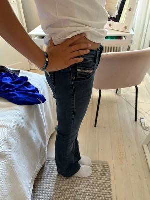 Mörkblå bootcut jeans DIESEL - Säljer ett par mörkblå bootcut jeans med låg midja och klassiska fem fickor. Jeansen har en snygg tvätt och subtila slitningar. Passar perfekt till en avslappnad stil och är tillverkade i stretchigt denimtyg för extra komfort. Diesel är långa i modellen