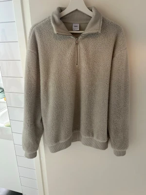 Beige fleece-stickad tröja med halvzip - Mysig beige stickad tröja i fleece med halv dragkedja framtill och ribbade muddar vid ärmslut och nederkant. Perfekt för kyliga dagar och har en avslappnad passform. Materialet är fluffigt och mjukt, vilket gör den extra skön att bära. Storlek L men passar M