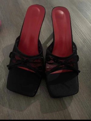 Svarta pumps med röd innersula - Snygga pumps med svart mesh och rosett framtill. Skorna har fyrkantig tå och en cool röd innersula som sticker ut. Perfekta för dig som vill ha något unikt och trendigt. Klackarna är öppna baktill och har en modern design.
