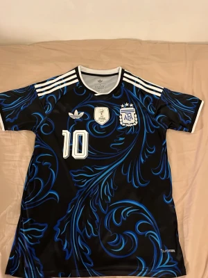 Argentina Messi matchtröja svart/blå - Snygg Argentina matchtröja från Adidas med Messi och nummer 10 på ryggen. Tröjan är svart med blått mönster, vita detaljer och tre vita ränder på axlarna. Korta ärmar och officiella klubbmärken framtill. Perfekt för fotbollsfans!