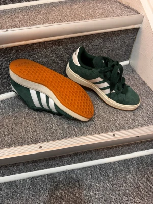 Adidas gröna sneakers med vita ränder - Mörkgröna Adidas sneakers i mocka med klassiska vita ränder på sidorna och vit hälflik med Adidas-logga. Skorna har en retroinspirerad siluett, tjock vit mellansula och gummisula i orange. Perfekta för dig som gillar streetwear och sportig stil.