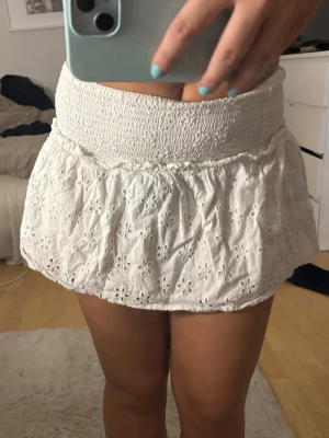 Vit kort kjol med broderade blommor - Supersöt vit kort kjol med bred smock i midjan och broderade blommor över hela tyget. Har haft denna kjol länge men inte andvänd på länge. Jätte söt till sommaren. Passar en storlek xs/s/m då den e väldigt stretchig. Köpt för 250 kr.