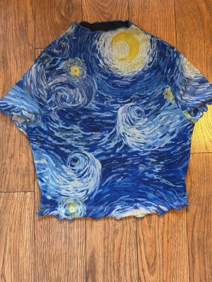 Blå topp med Van Gogh-motiv - Unik topp med korta ärmar och tryck inspirerat av Van Goghs 'Stjärnenatt'. Toppen har vågiga kanter och är i olika blå toner med inslag av gult och vitt. Materialet känns lätt och stretchigt, perfekt för dig som vill sticka ut.