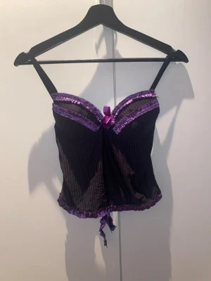 Svart och lila bustier med rosett - Snygg bustier i svart tyg med tunna vertikala ränder och lila spetsdetaljer. Dekorerad med en lila rosett framtill och volangkant både upptill och nedtill. Justerbara axelband och snörning nedtill för extra detalj. Perfekt för dig som vill ha något unikt och lekfullt.
