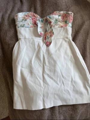 Vit axelbandslös klänning med blommor - Söt vit kortklänning med axelbandslös design och blommigt mönster upptill. Klänningen har en knytrosett framtill och är perfekt för varma dagar. Materialet känns som bomull eller linne och ger en fräsch och somrig vibe.