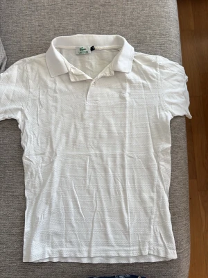 Vit randig pikétröja från Lacoste - Klassisk vit pikétröja från Lacoste med diskreta horisontella ränder och liten broderad logga på bröstet. Tröjan har krage, korta ärmar och knäppning framtill. Perfekt till jeans eller shorts för en clean look. Lite märke efter vikningen av kragen, därav bra pris.