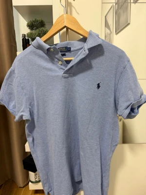 Blå pikétröja från Polo Ralph Lauren - Säljer en ljusblå pikétröja från Polo Ralph Lauren i slim fit-modell. Tröjan har korta ärmar, klassisk krage och knappar vid halsen. Ikoniska loggan broderad på bröstet. Perfekt för en clean och stilren look.