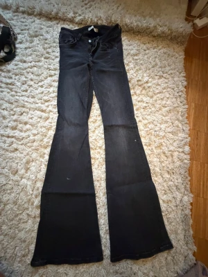 Svarta bootcut jeans med låg midja - Säljer ett par svarta bootcut jeans med låg midja och utsvängda ben. Jeansen har klassisk femficksdesign och knappgylf. Perfekta för dig som gillar retrovibbar och vill ha ett par riktigt snygga jeans med breda ben.