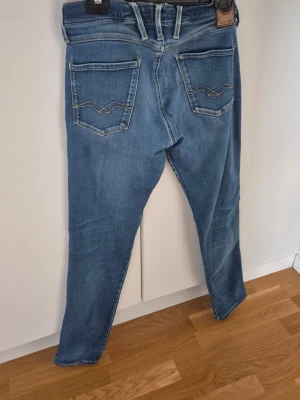 Replay Anbass Jeans - Säljer ett par blå slim fit jeans med snygga slitningar framtill och dekorativa bakfickor med broderade detaljer. Jeansen har klassisk femficksmodell, normalhög midja och dragkedjegylf. Perfekta för en avslappnad och trendig look.