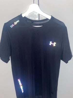 Svart tränings-tshirt Under Armour - Svart funktions-tshirt från Under Armour med reflekterande logga på bröstet och textdetaljer på axel och nederkant. Tillverkad i ett lätt och snabbtorkande material, perfekt för löpning eller gym. Klassisk rund hals och korta ärmar.