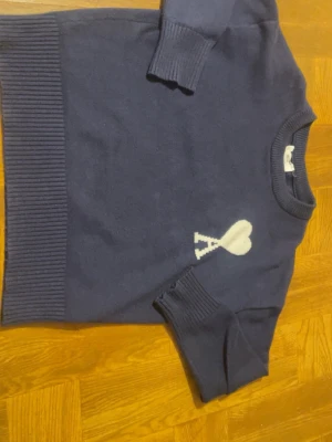 Mörkblå stickad tröja med hjärtdetalj - Mörkblå stickad tröja med korta ärmar och ribbade muddar. På bröstet finns en vit hjärtdetalj med bokstaven A under. Tröjan har rund halsringning och är gjord i ett mjukt material som känns skönt mot huden.