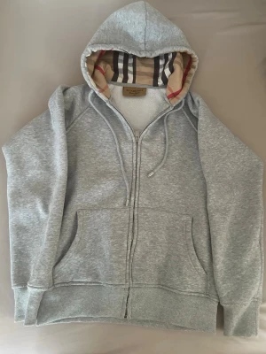 Burberry zip hoodie - Snygg grå zip hoodie från Burberry med klassiskt rutigt foder i huvan. Hoodien har dragsko i huvan, två stora fickor framtill och ribbade muddar vid ärmar och nederkant. Perfekt för en avslappnad och trendig look.