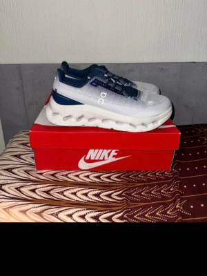 Vita och marinblå sneakers från On - Snygga sneakers från On i vitt och marinblått med mesh-överdel och unika, vågformade sulor. Skorna har blå detaljer och vita snören med små turkosa accenter. Perfekta för dig som gillar sportig och modern stil.