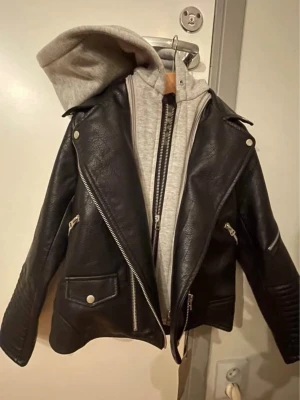 Svart skinnjacka med hoodie från Zara - Snygg svart skinnjacka från Zara med grå hoodie under. Jackan har flera dragkedjor, breda slag och coola detaljer på ärmarna. Perfekt för dig som vill ha en edgy look med streetkänsla. Hoodie och jacka sitter ihop för extra stilpoäng.