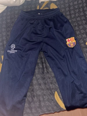 Blå FC Barcelona träningsbyxor - Mörkblå träningsbyxor med FC Barcelona-märke på höger ben och Champions League-logga på vänster ben. Byxorna har resår i midjan och är gjorda i ett lätt, sportigt material som passar perfekt till fotbollsträning eller chill. Snygg och bekväm passform.