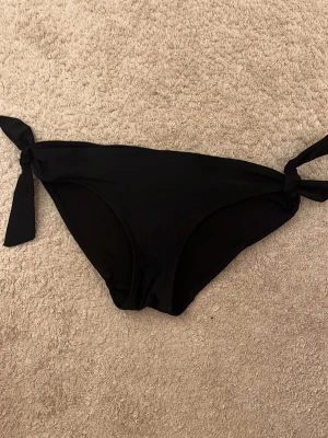 Svarta bikinitrosor med knyt - Snygga svarta bikinitrosor med knyt i sidorna. Klassisk modell som passar perfekt till sommaren och strandhäng. Trosorna har en enkel och stilren design som är lätt att matcha med olika bikiniöverdelar.