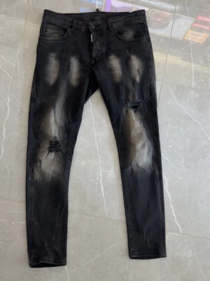 Äkta Svarta Dsquared 2  jeans - Säljer ett par svarta skinny jeans med slitningar och blekta detaljer på benen. Jeansen har klassisk femficksmodell och dragkedjegylf. Perfekta för dig som gillar en edgy och trendig stil.
