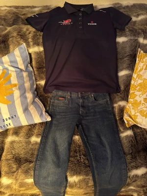 Blå jeans från Wrangler och piké från en collab av sail racing och redbull - Säljer ett par klassiska blå jeans från Wrangler med fem fickor och raka ben. Jeansen har en snygg tvätt och detaljerade sömmar. Perfekta för dig som gillar en tidlös och avslappnad stil. Och en piké från sail racing