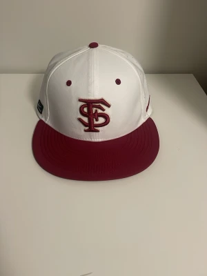 SnapBack keps - Snygg snapback keps i vitt med vinrött brätte och broderad logga framtill. Kepsen har en klassisk rak skärm och små ventilationshål bak. Perfekt för dig som vill ha en stilren och sportig look.