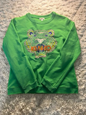 Grön Kenzo sweatshirt med tigertryck - Cool grön sweatshirt från Kenzo med det ikoniska broderade tigerhuvudet och färgglada detaljer framtill. Tröjan har rund halsringning och ribbade muddar vid ärmslut och nederkant. Perfekt statement-plagg för dig som vill sticka ut.