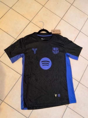 Svart och lila FC Barcelona matchtröja - Snygg svart och blå FC Barcelona matchtröja med klubbmärke och Spotify-logga på bröstet. Tröjan har korta ärmar, sportig passform och är tillverkad i ett lätt, ventilerande material. Perfekt för fotbollsträning eller att bära till vardags.
