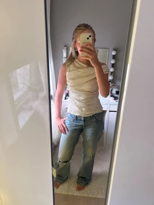 Beige omlott ärmlös topp - Snygg beige topp med omlott-detalj och ärmlös design. Toppen har en lätt glansig yta och sitter figurnära med en modern look. Perfekt att styla med jeans eller kjol för en trendig outfit.