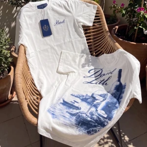 Vit t-shirt med blått tryck - Säljer en vit t-shirt med blått tryck framtill och stort grafiskt motiv på ryggen. T-shirten har rund hals och korta ärmar. Materialet är mjuk bomull och passformen ser ut att vara normal. Perfekt för dig som gillar stilrena plagg med en twist.