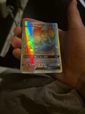 Pokémonkort Alolan Golem GX holo - Säljer ett Pokémonkort av typen Alolan Golem GX med holografisk yta. Kortet har färger i blått, orange och gult och visar en kraftfull Pokémon med attacker som Hammer In och Heavy Rock GX. Perfekt för samlare eller Pokémonfans.