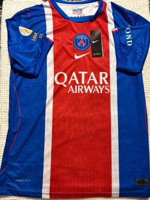 PSG matchtröja blå/röd Nike 2026/2027 - Snygg PSG matchtröja i blått och rött med vita detaljer. Tröjan har klubbmärke, Nike-logga och Qatar Airways-tryck på bröstet. Tillverkad i lätt och ventilerande material med Aero-Fit-teknologi. Perfekt för fotboll eller träning.köpt utomlands