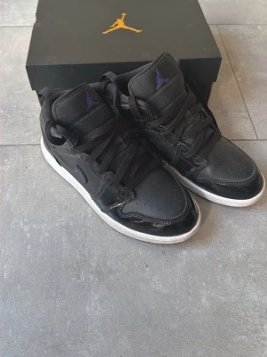 Svarta Air Jordan 1 Mid sneakers - Snygga svarta Air Jordan 1 Mid sneakers med glansiga detaljer och klassisk Nike swoosh på sidan. Skorna har svart ovandel i läder och textil, svart snörning och lila Jumpman-logga på plösen. Yttersulan är vit med grått mönster. Perfekta för dig som gillar streetwear. Mycket fint skick och väldigt sparsamt använda. Nypris 1100kr