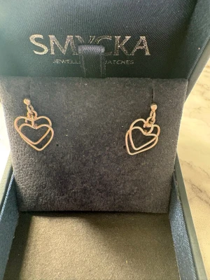 Silverörhängen med dubbla hjärtan - Säljer ett par stilrena örhängen i silverfärg med dubbla hjärtan i öppen design. Örhängena hänger i små kedjor och ger en modern och romantisk look. Perfekta för dig som gillar minimalistiska smycken med en twist.