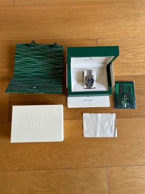 Klocka - Säljer en stilren klocka i silver med länkarmband och svart urtavla. Klockan kommer i originalbox med certifikat, putsduk och påse. Perfekt för dig som vill ha en klassisk och exklusiv accessoar.