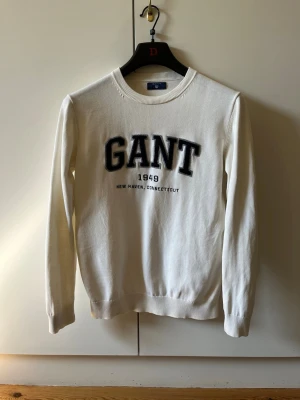 Vit tröja från GANT strl S - Klassisk vit långärmad tröja från GANT med svart broderat logotyp-tryck och texten '1949 New Haven, Connecticut' på bröstet. Rund halsringning och ribbade muddar. Har ett lagat hål, se sista bild. Material 100% bomull. Storlek S.
